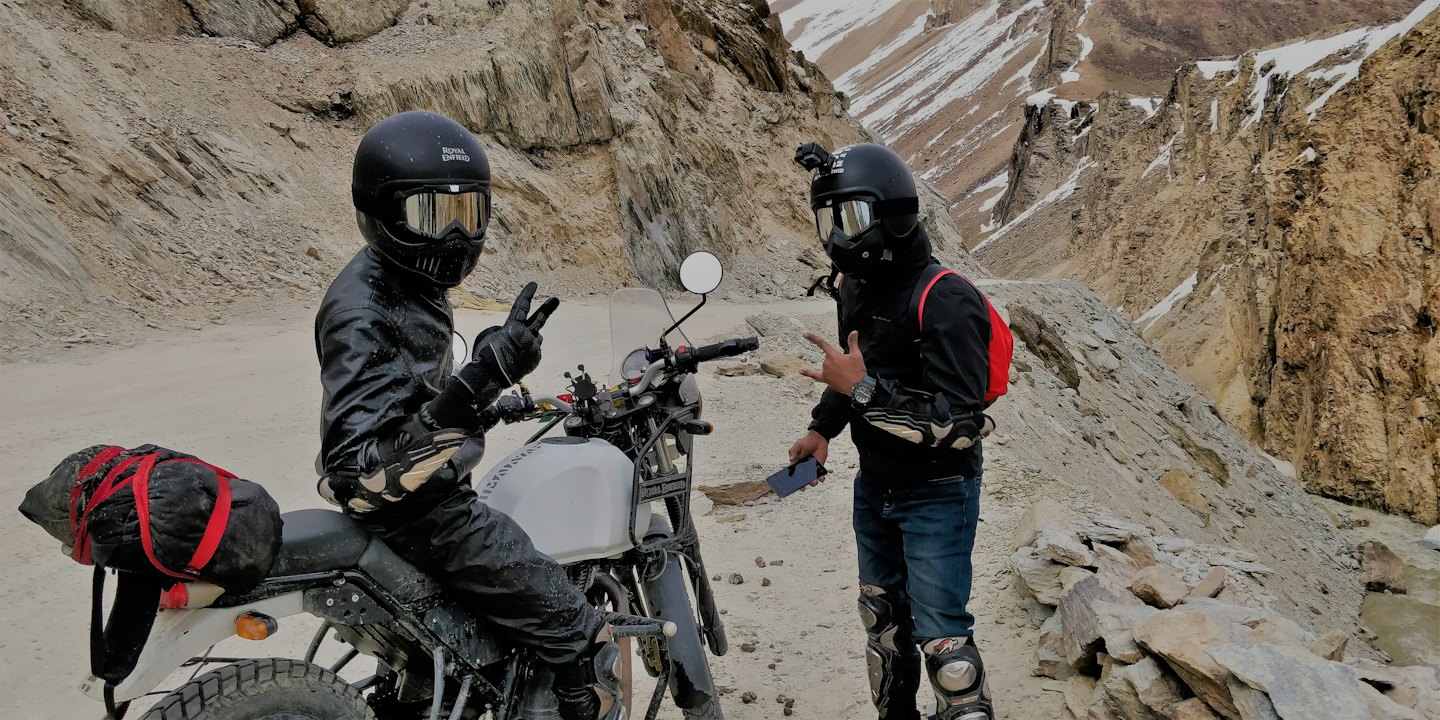 Two motorcyclists pose together on a rugged dirt mountain road.